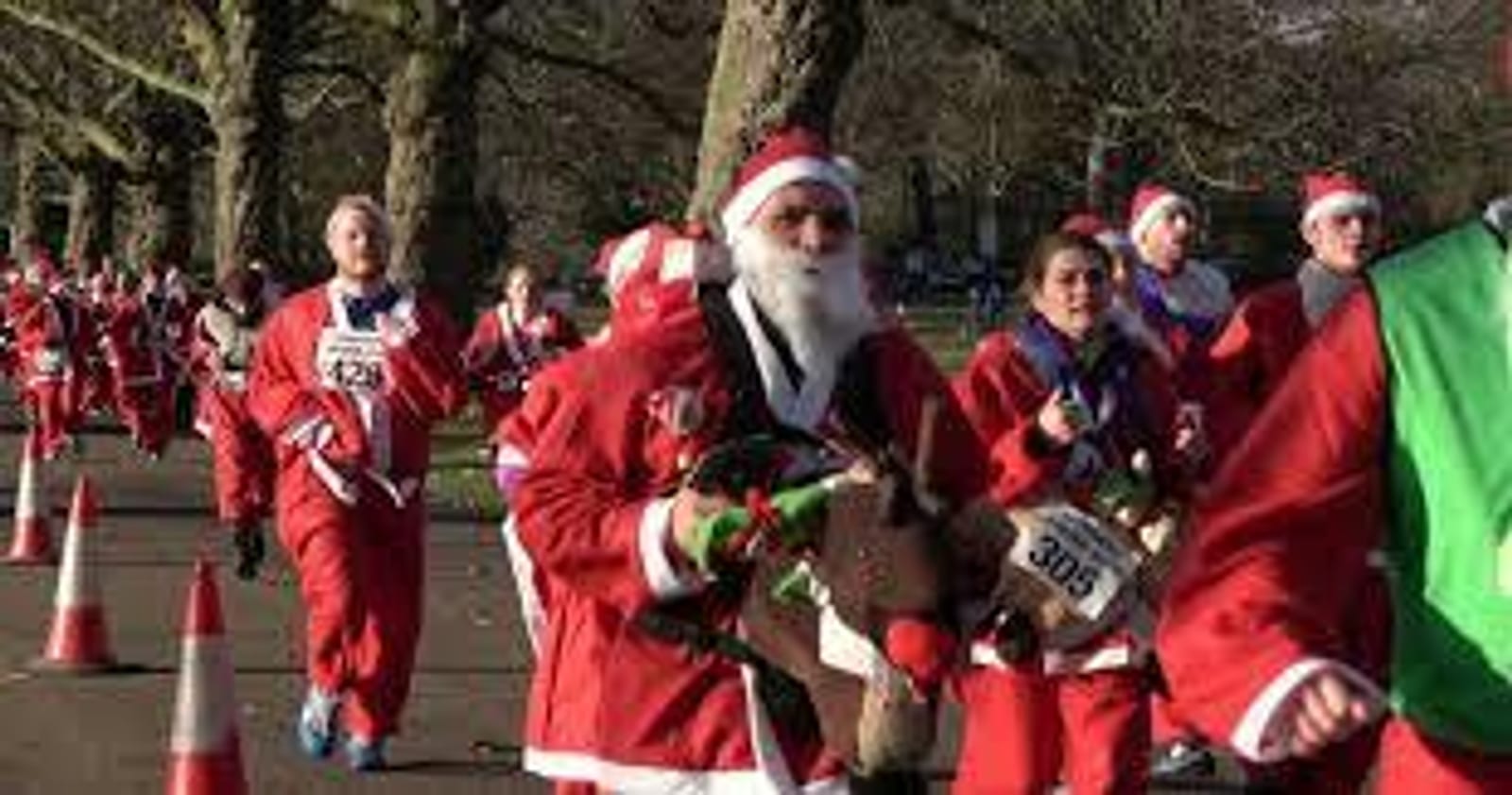 A group of people dressed in Santa Claus costumes participate in an outdoor fun run. One person in the foreground holds a stuffed reindeer. Orange cones line the path, with trees visible in the background. The atmosphere appears festive and lively.