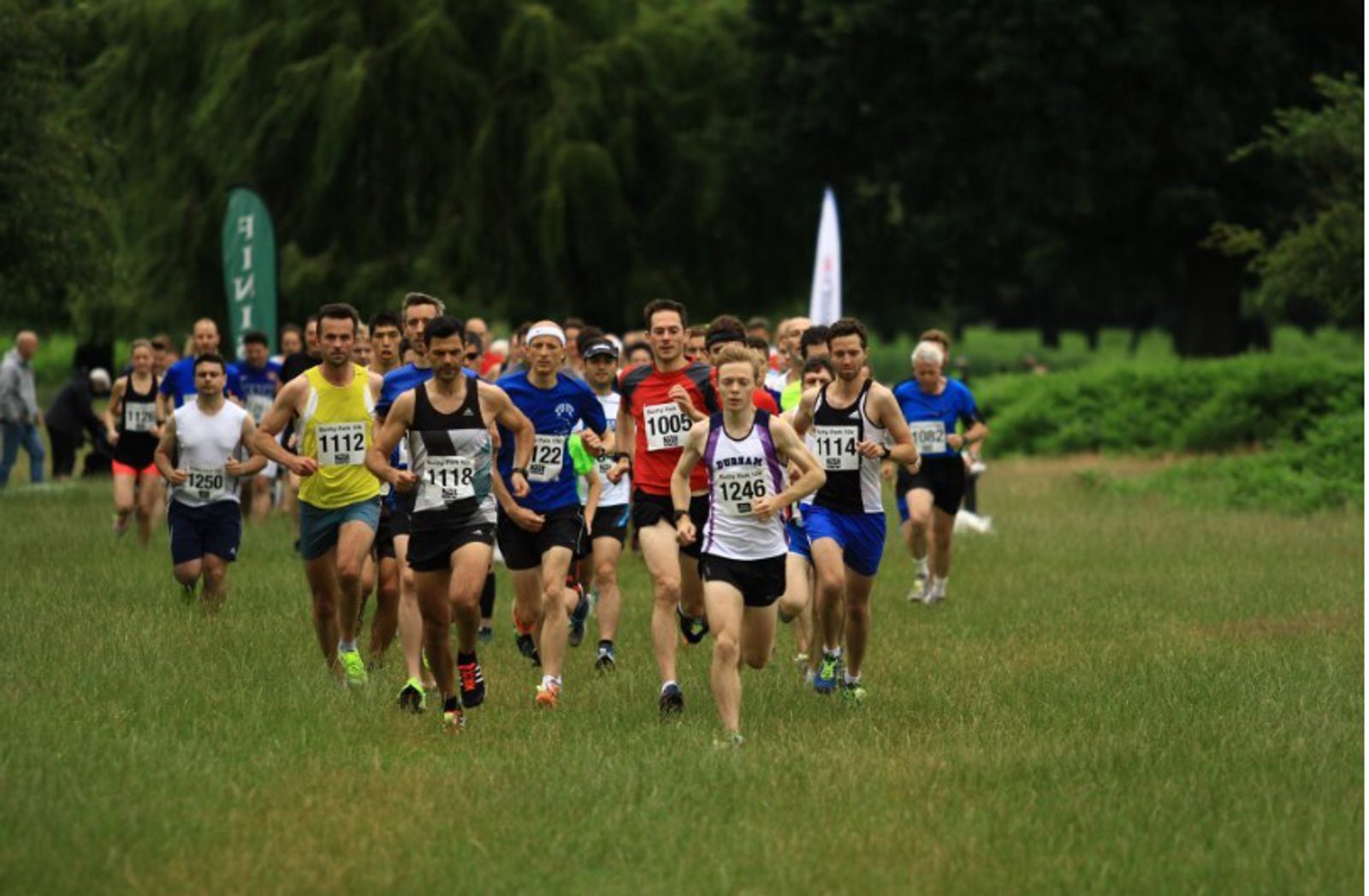 A group of runners participating in a race on a grassy field. The runners wear various athletic outfits and numbered bibs, with one runner in a white and black outfit leading the pack. Trees and other participants are visible in the background.