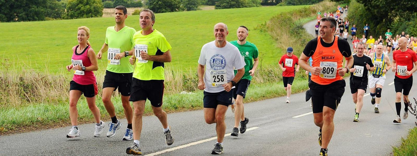 A group of runners participating in a road race on a countryside road. The participants are wearing numbered race bibs and various brightly colored athletic clothing. In the background, there are more runners, green fields, and trees along the sides of the road.