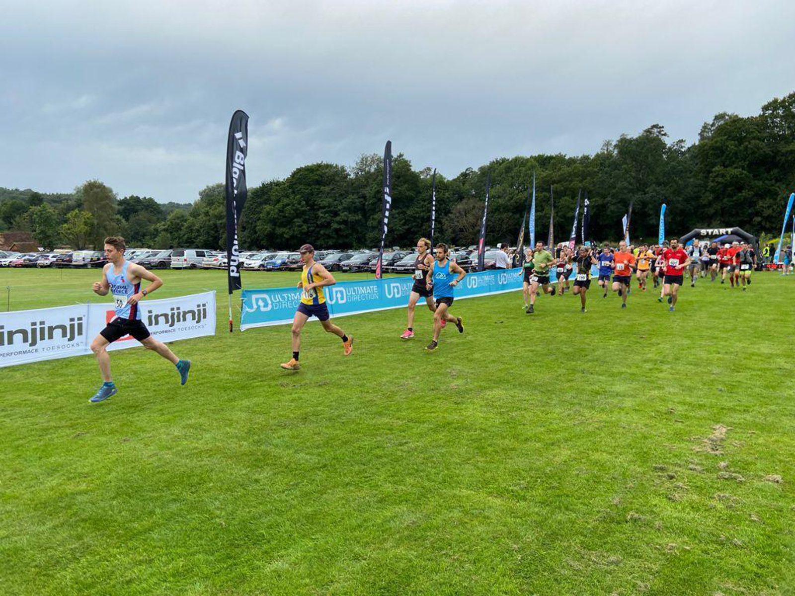 A group of runners competing in a race on a grass field, with several runners wearing colorful athletic clothing. Banners and flags with sponsor logos line the course, while in the background, trees and a cloudy sky can be seen. Spectators are visible on the right.