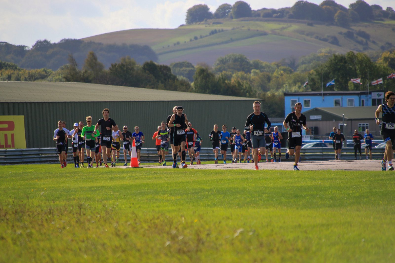 A group of runners participating in a race on a sunny day, with lush green fields and rolling hills in the background. Some participants are wearing numbers on their chests, and there are cones along the path guiding the runners.