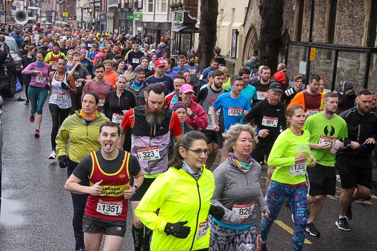 A large group of runners participates in a road race on a city street. They wear colorful athletic clothing, some with bib numbers displayed. One runner in the foreground playfully sticks out his tongue, while others focus ahead. Crowds line the street in the background.