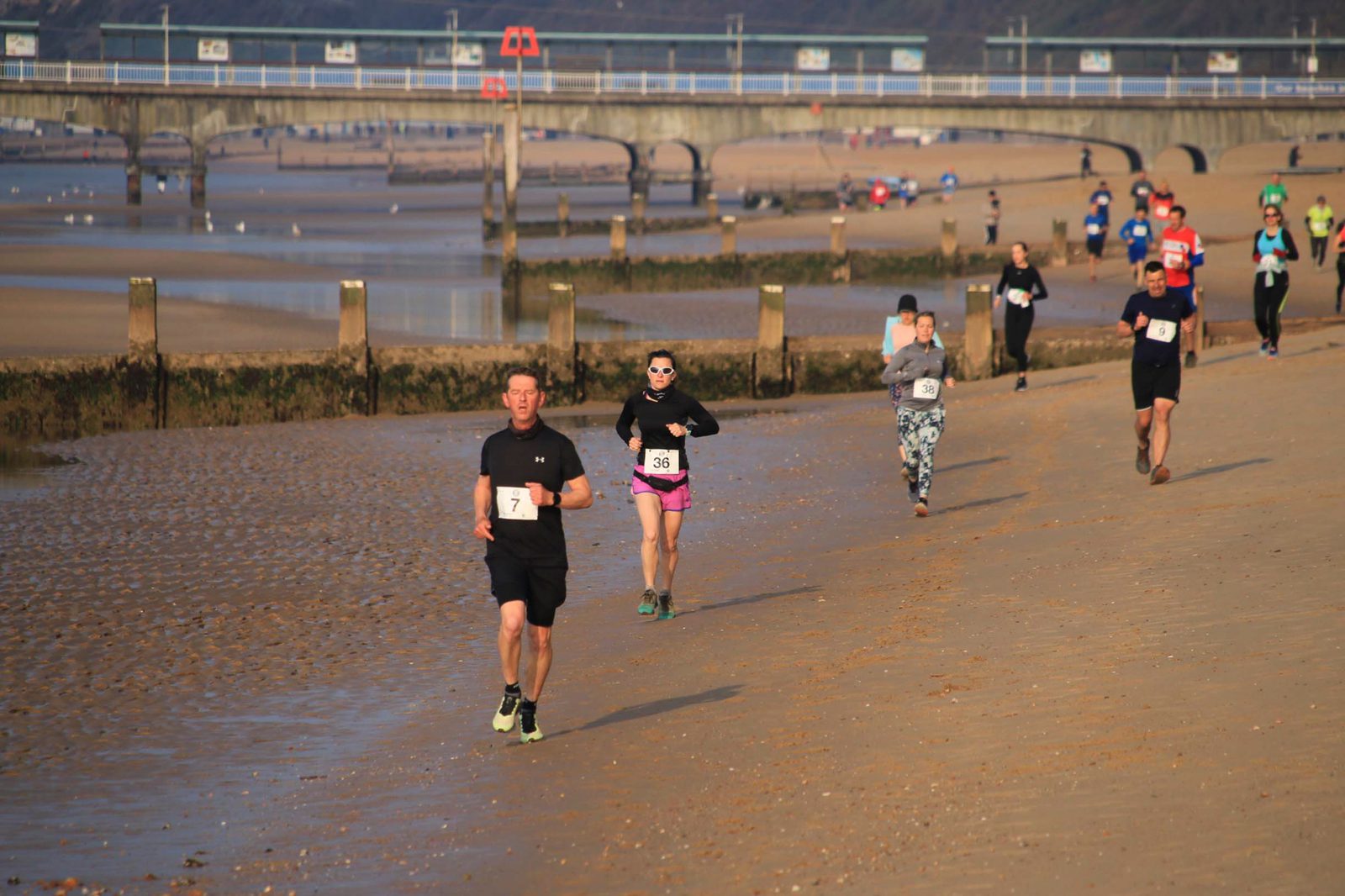 A group of runners participating in a race on a sandy beach. The tide is low, revealing wet sand, and a pier extends into the water in the background. Several runners are wearing numbered bibs, with some dressed in athletic attire including shorts, leggings, and sunglasses.