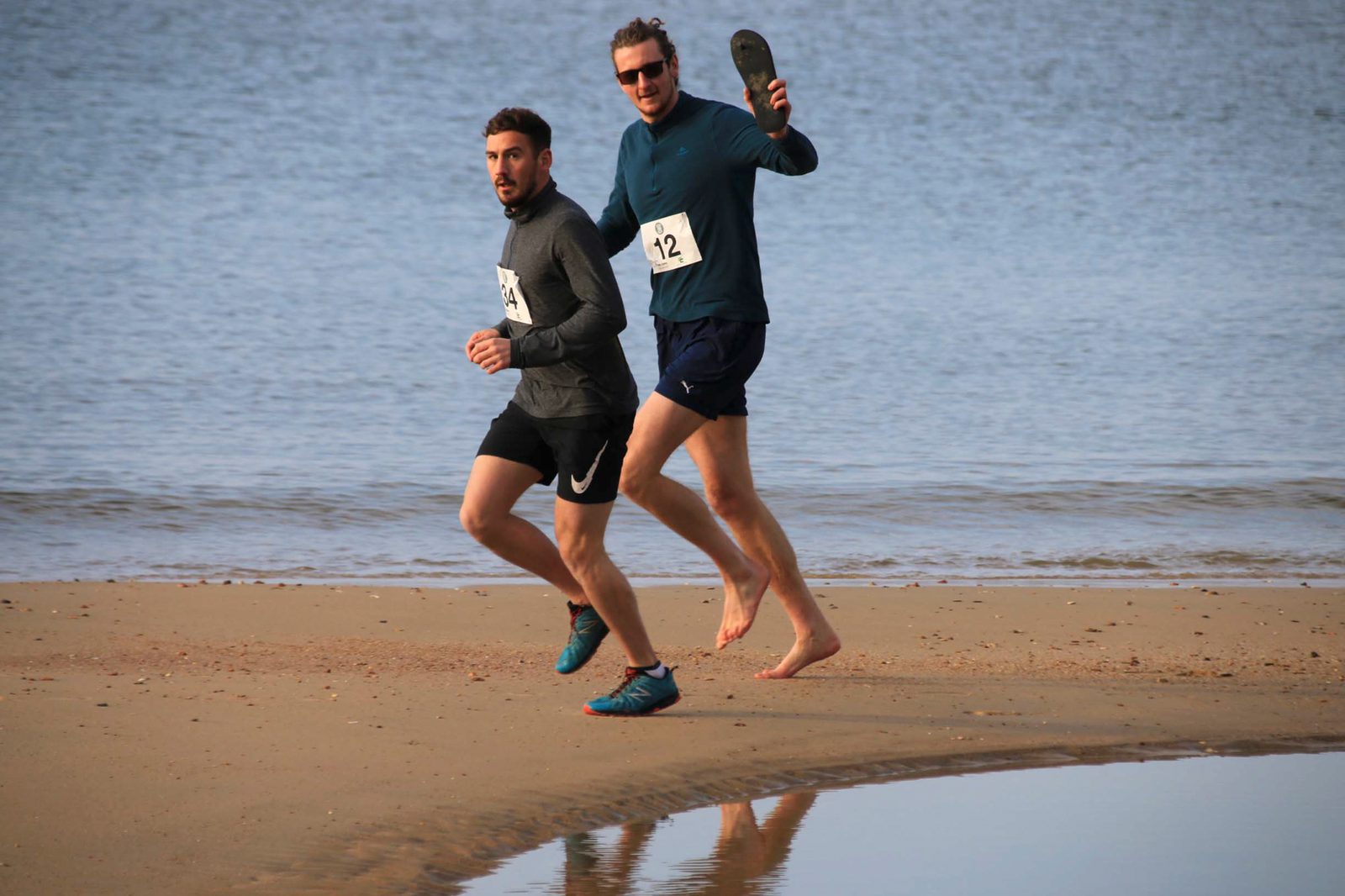 Two men are running on a sandy beach near the water. Both are wearing running gear with race numbers pinned to their shirts. One of the men is barefoot and holding a sandal in one hand, while the other man is wearing running shoes. They appear to be in motion.