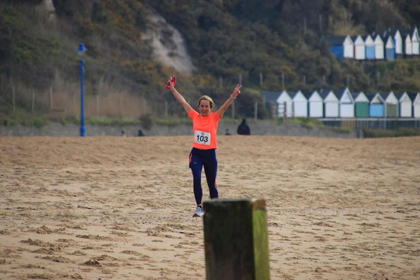 A runner wearing an orange shirt and blue leggings, with a race number 103, celebrates with raised arms as they run on a sandy beach. Beach huts and a hill are visible in the background.