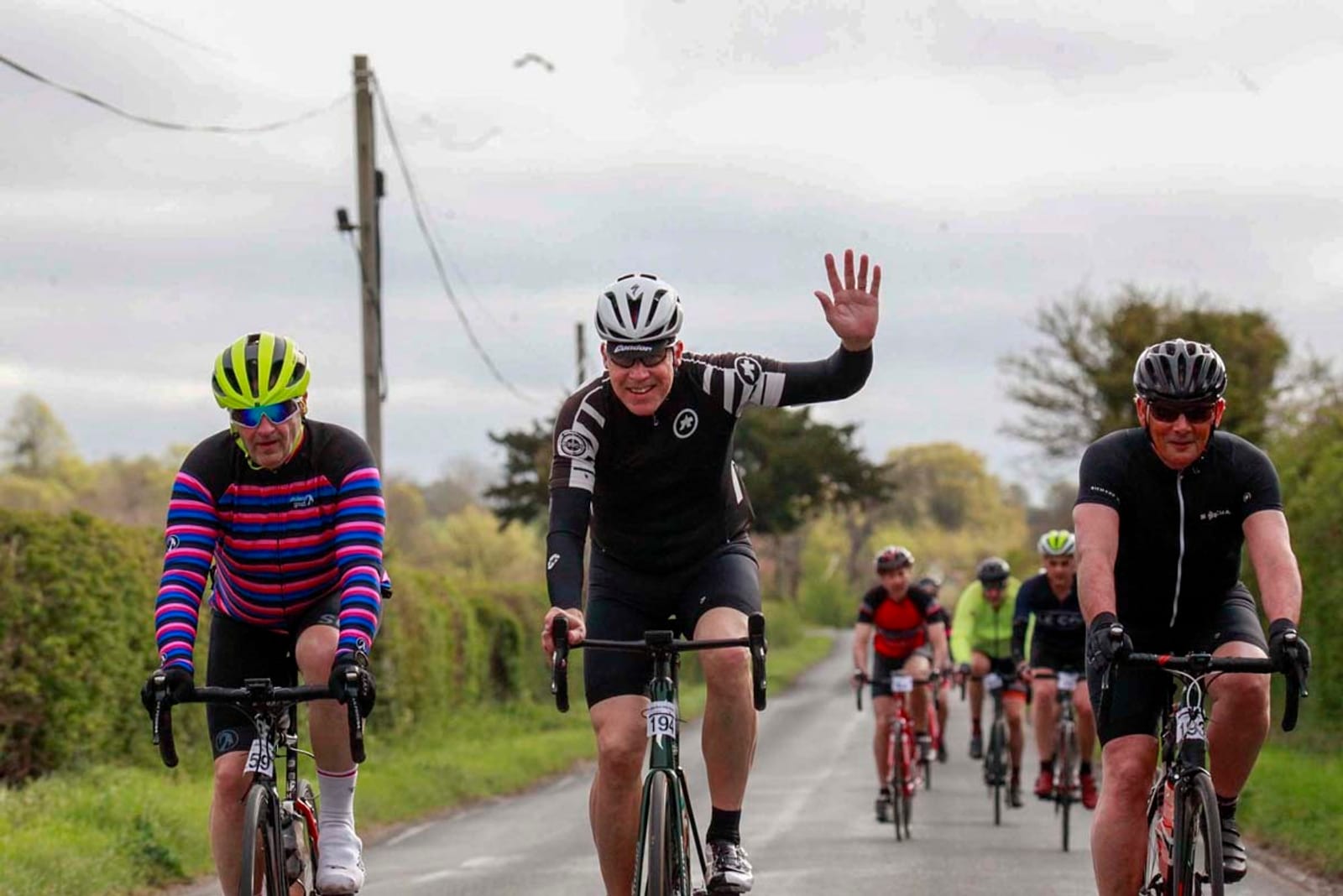 A group of cyclists ride on a rural road. The cyclist in the center wears a black and white outfit, helmet, and is waving with a smile. The cyclist on the left wears a colorful striped jersey and bright green helmet, while the one on the right wears black attire and a helmet.