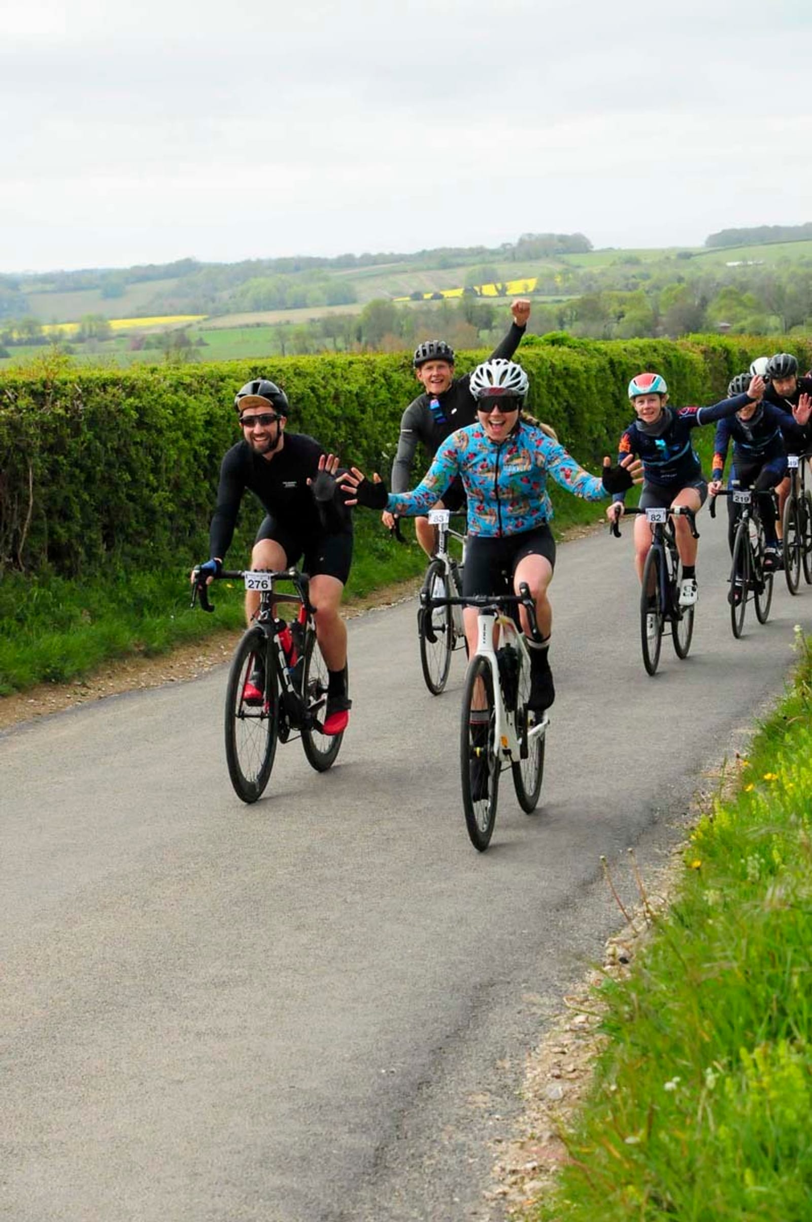 A group of cyclists rides along a countryside road on a cloudy day. The cyclists, wearing helmets and cycling attire, appear happy and are waving. The background includes green hedges, fields, and slightly hilly terrain in the distance.