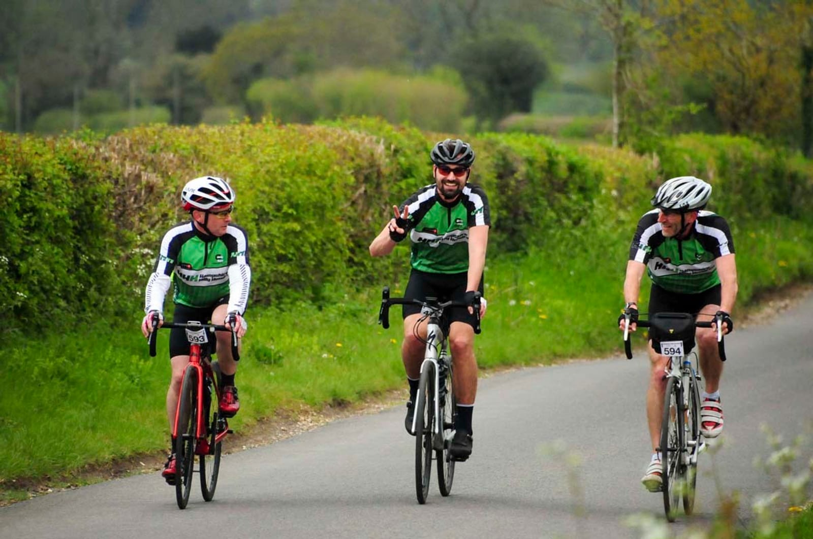 Three cyclists in green and black jerseys ride along a country road lined with green bushes and trees. The middle rider smiles and gives a peace sign while the others ride alongside, enjoying the scenery and pleasant weather.