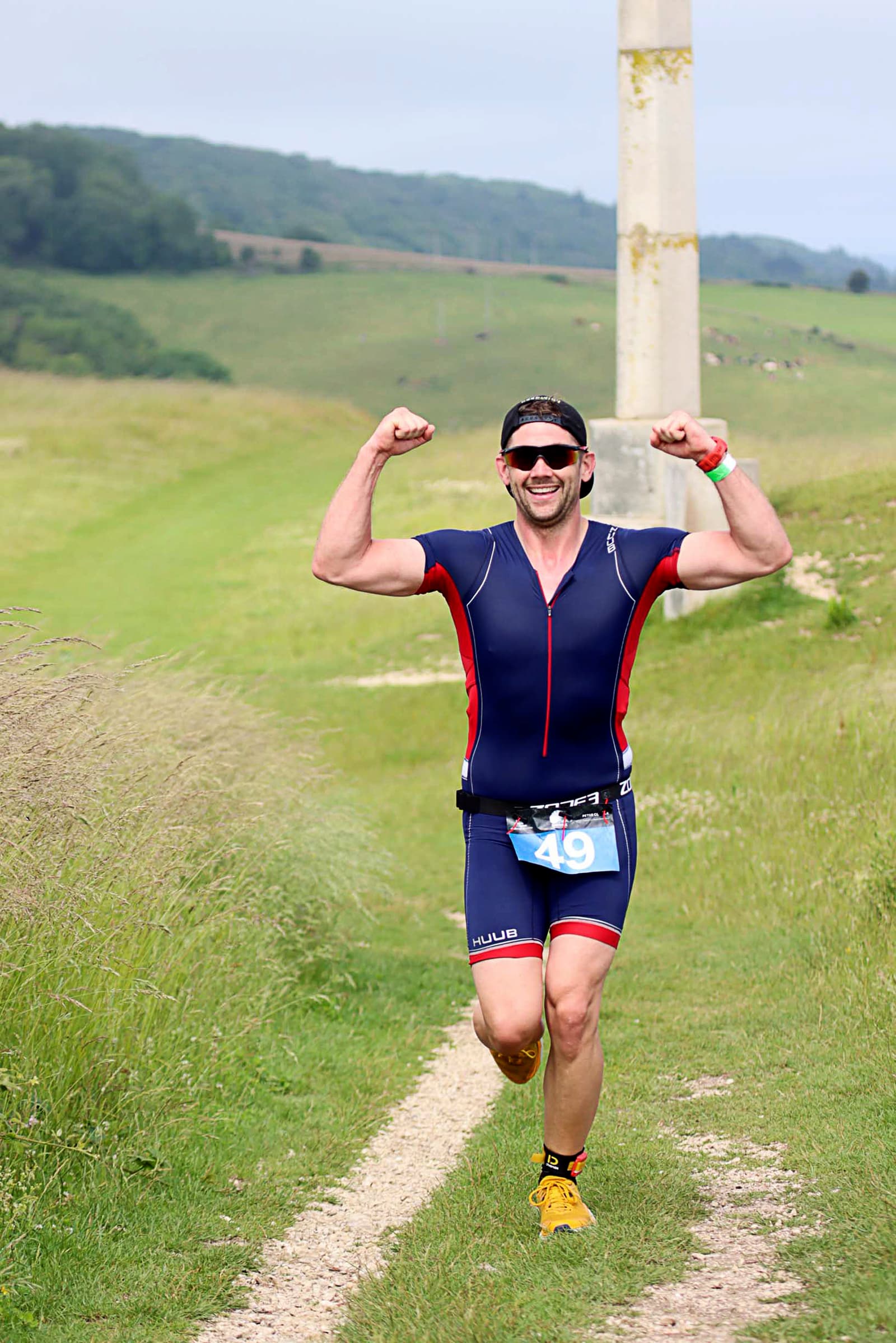 A male athlete wearing a navy blue triathlon suit and black sunglasses is running on a grassy trail with his arms flexed in a victory pose. He is smiling and has race number 49 attached to his suit. The background features rolling hills and a clouded sky.