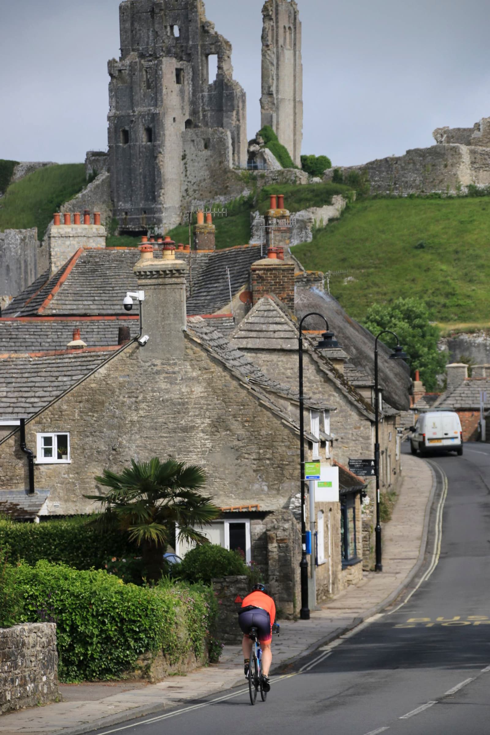 A person in a red jersey and black shorts rides a bicycle on a narrow, gently curving street lined with stone houses. In the distance, an ancient stone castle ruin sits atop a grassy hill under a cloudy sky.