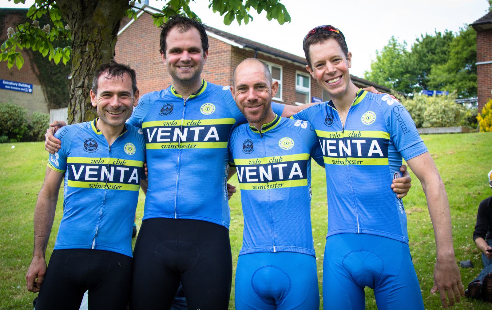 Four cyclists wearing matching blue and yellow "Venta" cycling jerseys stand outdoors with their arms around each other, smiling at the camera. They are in front of a brick building and a green, grassy area with trees.