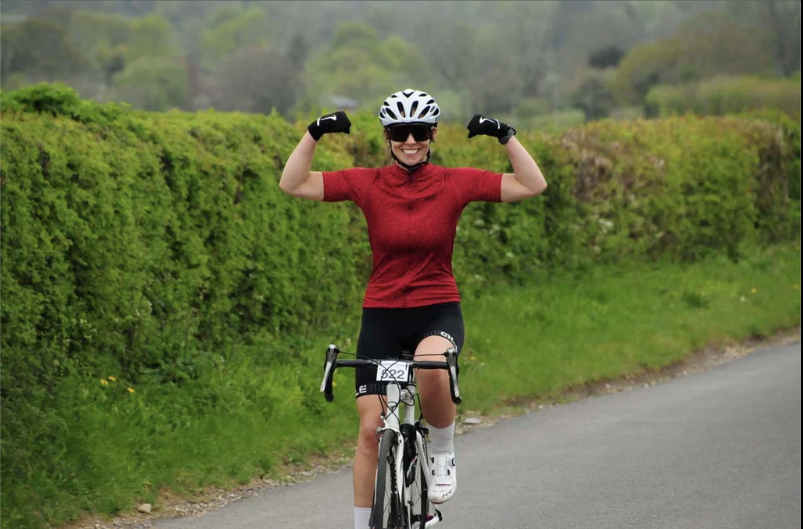 A cyclist wearing a red shirt, black shorts, and a white helmet is riding on a road lined with green bushes. The cyclist is smiling and flexing both arms, showing a gesture of strength or triumph. The background includes a landscape of trees and a cloudy sky.