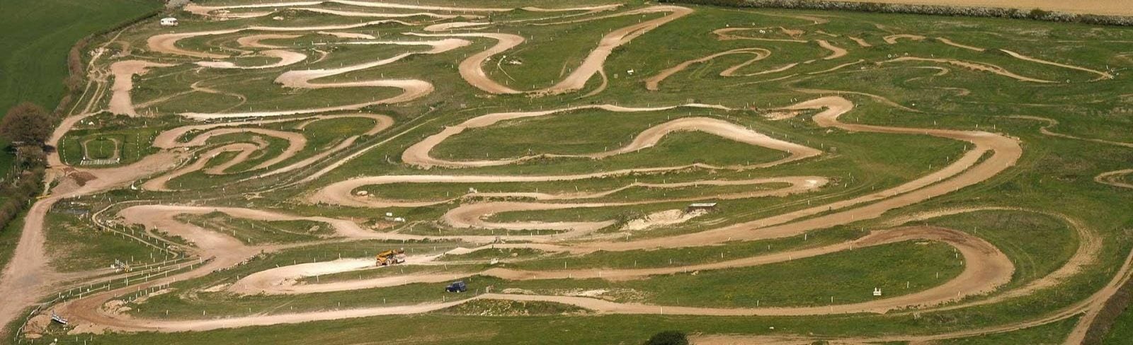 Aerial view of an extensive motocross track featuring numerous dirt paths winding and twisting through green fields. The track includes sharp turns, steep inclines, and various obstacles designed for off-road motorcycle racing.