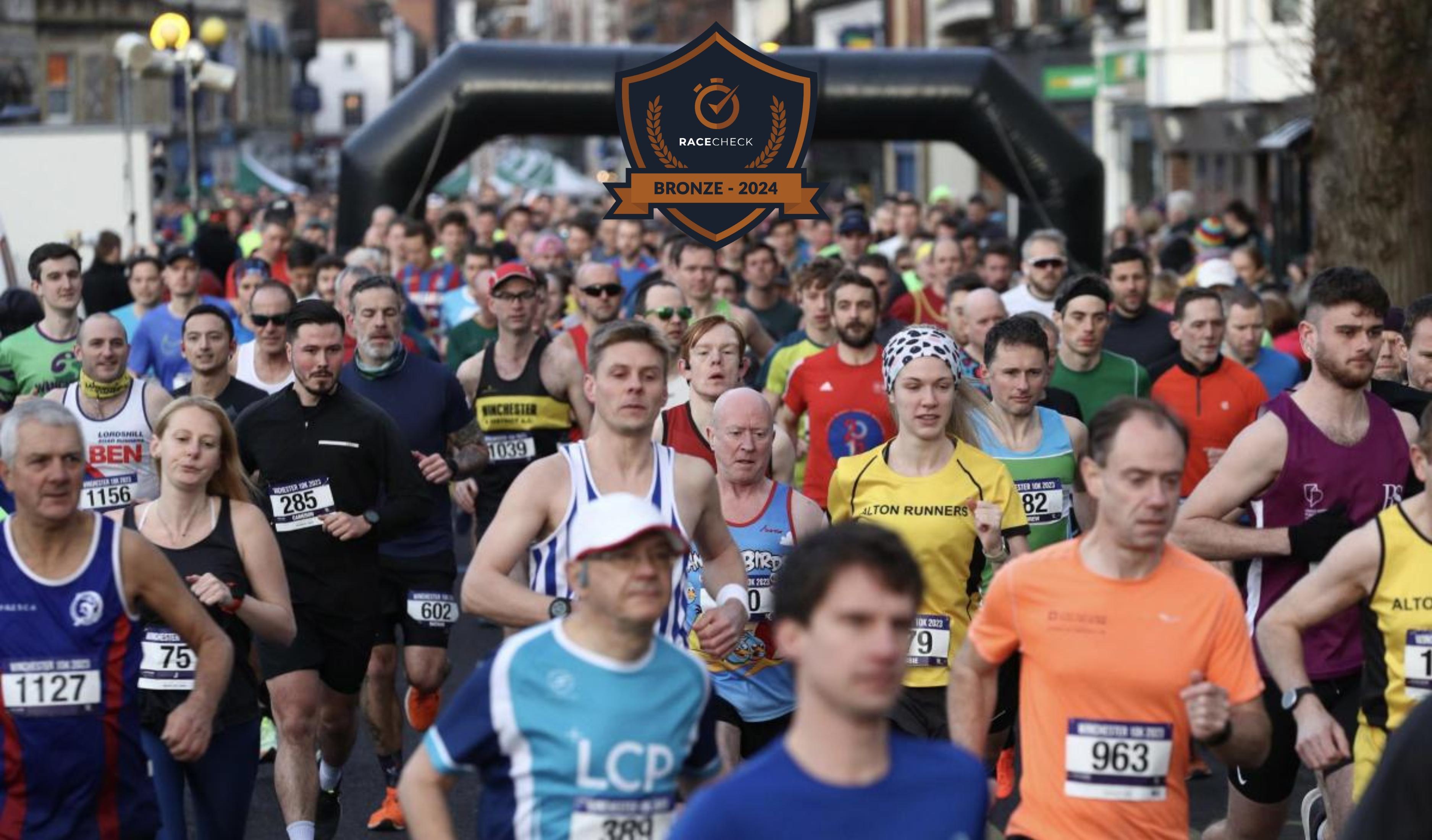 A large group of runners participating in a street race. They are wearing various athletic gear and running in a bustling urban environment. A banner above them reads "Bronze - 2024.