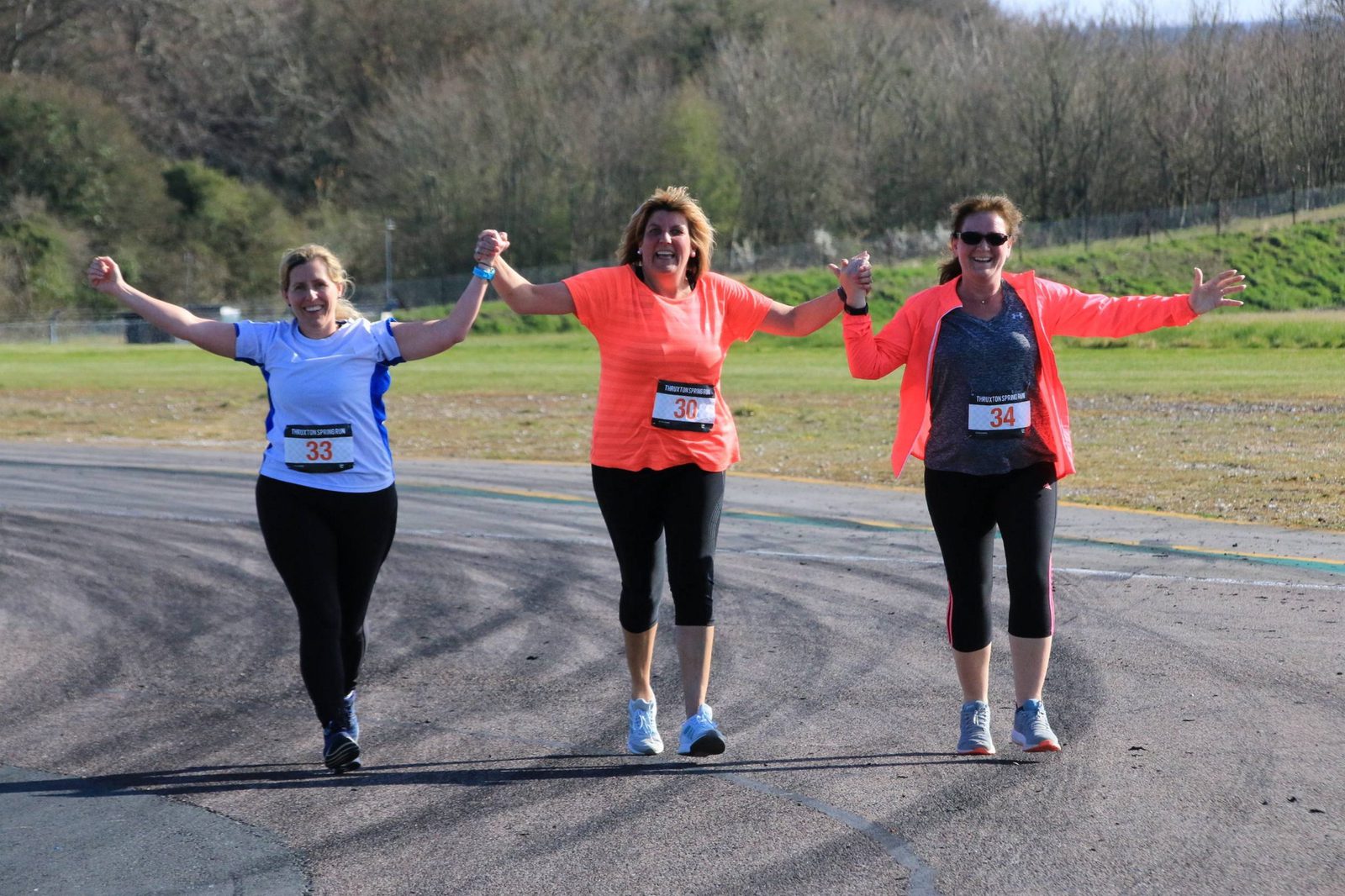 Three women in athletic wear, each with a race number (33, 30, 34), walk hand-in-hand on a paved track, smiling and holding their arms up in celebration. Trees and grassy areas are visible in the background.