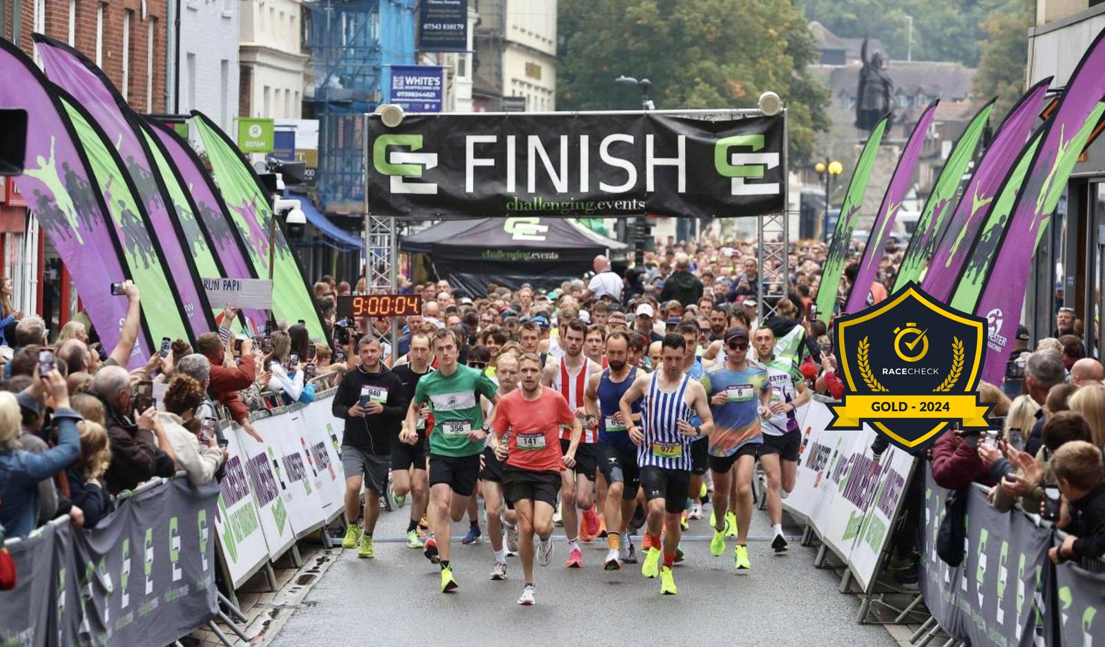 Runners start a race on a city street, passing under a "Finish" banner. Spectators line the sides, and a digital timer shows 9:00:04. A "Race Check Gold - 2024" badge is displayed. The atmosphere is lively and energetic.