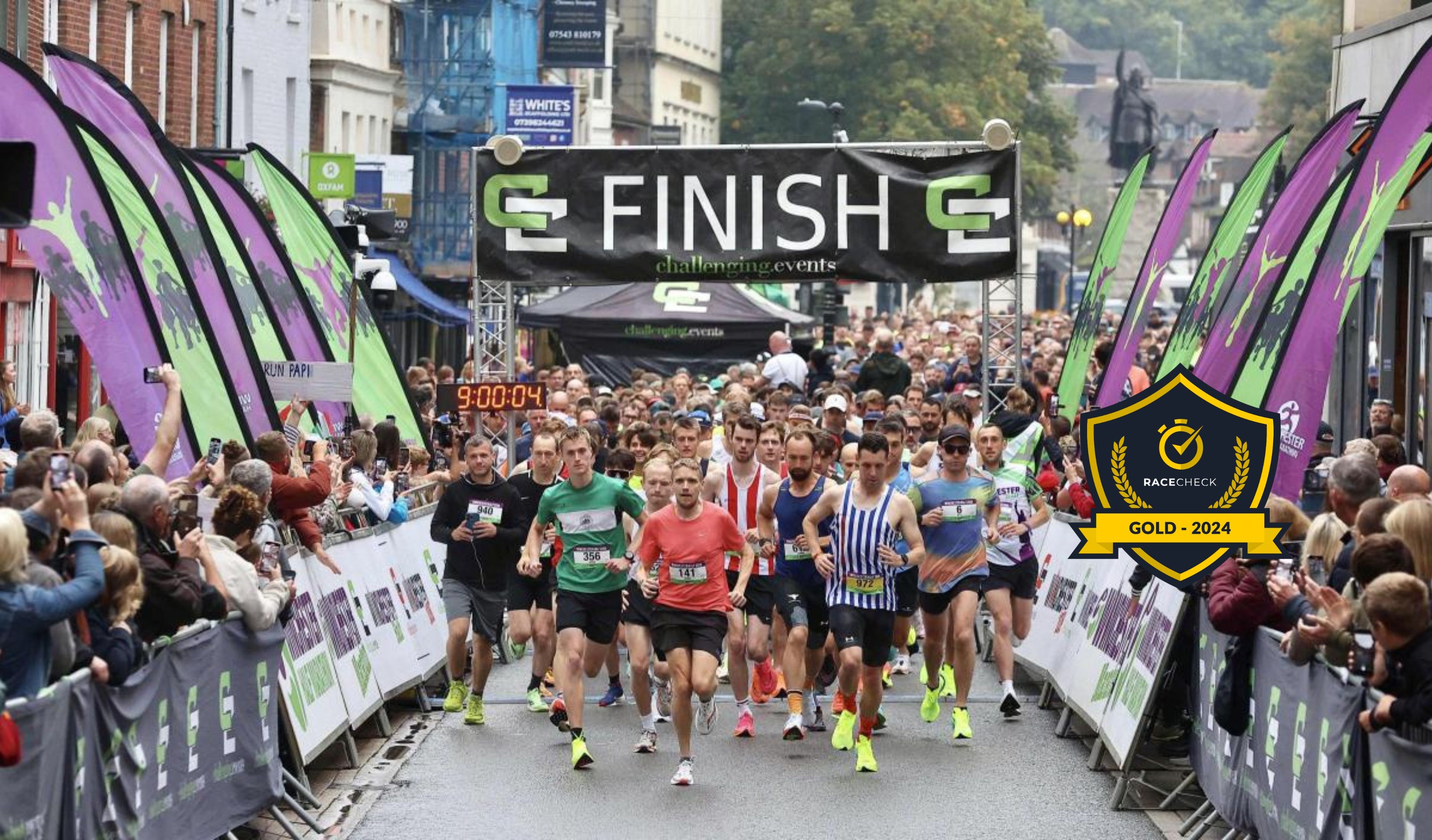Runners start a race on a city street, passing under a "Finish" banner. Spectators line the sides, and a digital timer shows 9:00:04. A "Race Check Gold - 2024" badge is displayed. The atmosphere is lively and energetic.