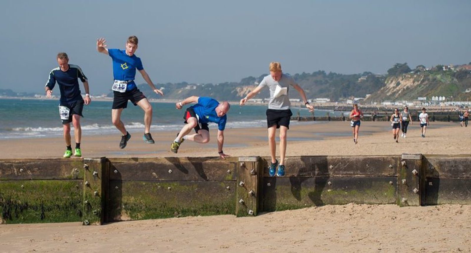 Four runners in athletic attire jump over a wooden barrier on a sandy beach during a race. Other participants can be seen running in the background, with the ocean and a scenic coastline visible. The sky is clear and sunny.
