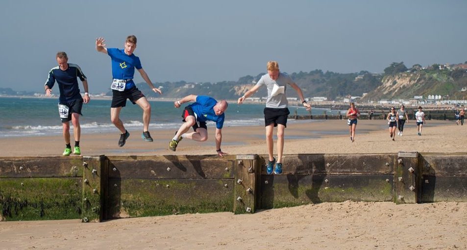 Four runners in athletic attire jump over a wooden barrier on a sandy beach during a race. Other participants can be seen running in the background, with the ocean and a scenic coastline visible. The sky is clear and sunny.