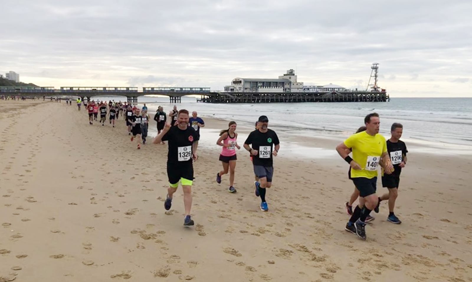 A group of runners in athletic wear participate in a race along a sandy beach. A pier extends over the ocean in the background under a cloudy sky. Footprints in the sand trail behind the runners as they advance. The scene exudes energy and determination.