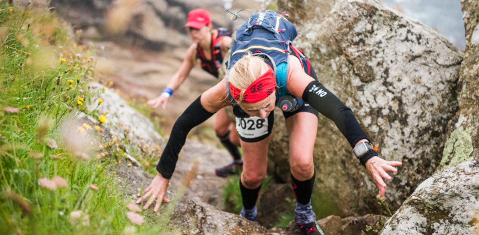Two trail runners wearing race bibs navigate a rocky ascent. The leading runner, a woman with a red headband and black arm sleeves, climbs with both hands on the rocks. The second runner, also wearing a headband, follows closely. Grass and wildflowers are visible.