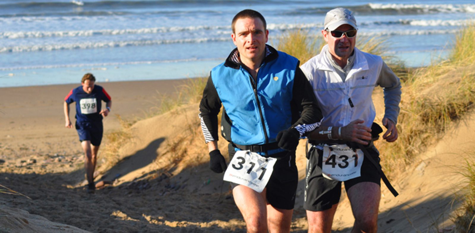 Three runners are participating in a race on a sandy beach with the ocean in the background. The two closest runners are wearing numbered bibs (311 and 431) and are ahead of the third runner, who is further back, wearing bib 398. The day appears to be sunny.