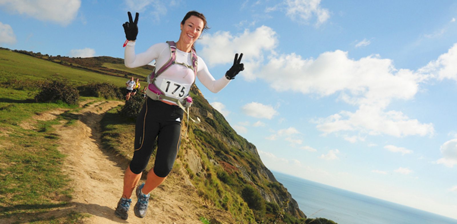 A woman wearing athletic clothing and a race number "175" runs on a trail near a hillside with the ocean in the background. She is smiling and making peace signs with both hands. The weather is clear with a blue sky and a few clouds.