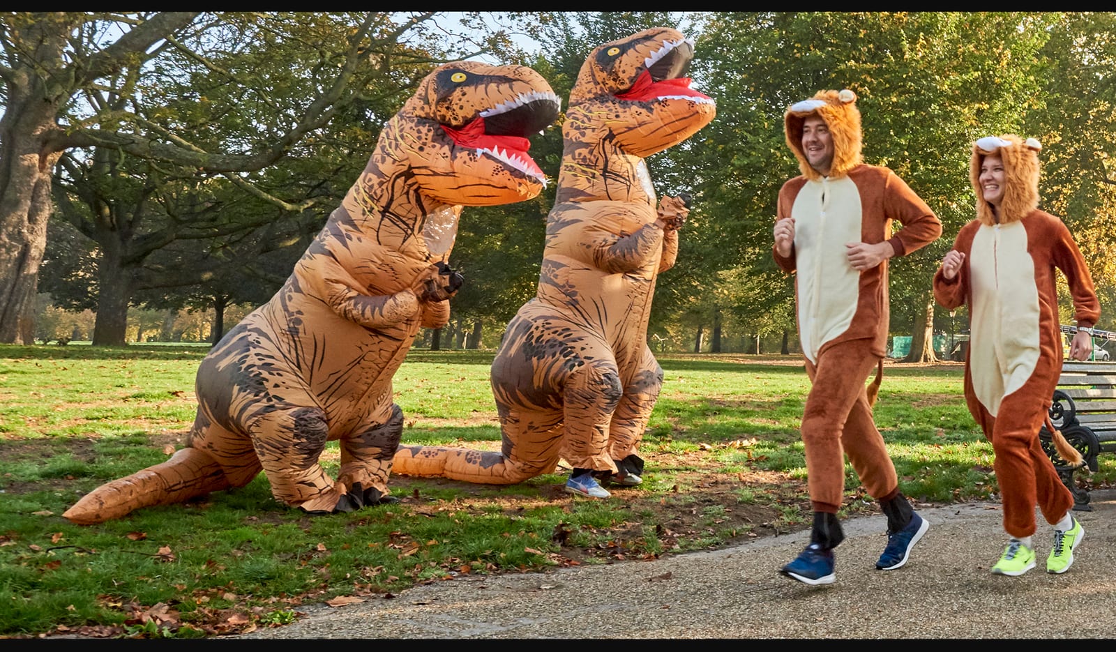 Two people in lion costumes run in a park on a paved pathway, while two individuals in inflatable T-Rex dinosaur costumes stand nearby. The background features trees and green grass under a clear sky. The scene is vibrant and playful.