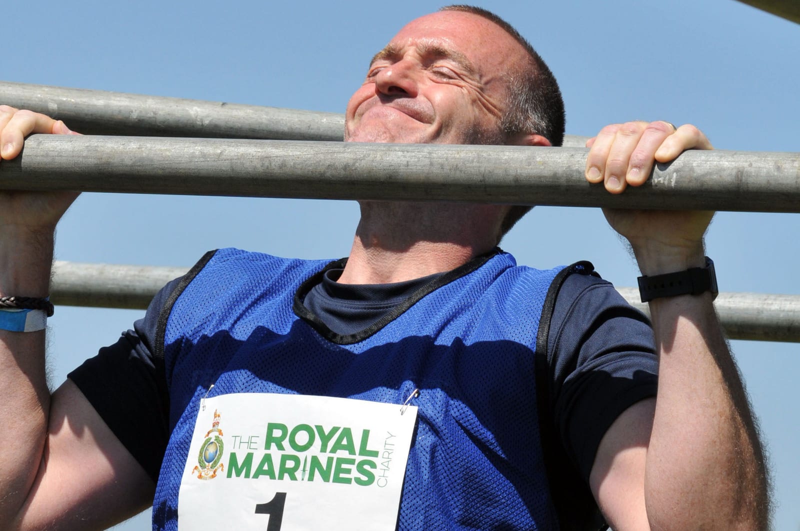 A man in a blue jersey performs a pull-up on a metal bar during an outdoor activity. He is wearing a race number bib with "The Royal Marines Charity" printed on it. The sky is clear and blue.
