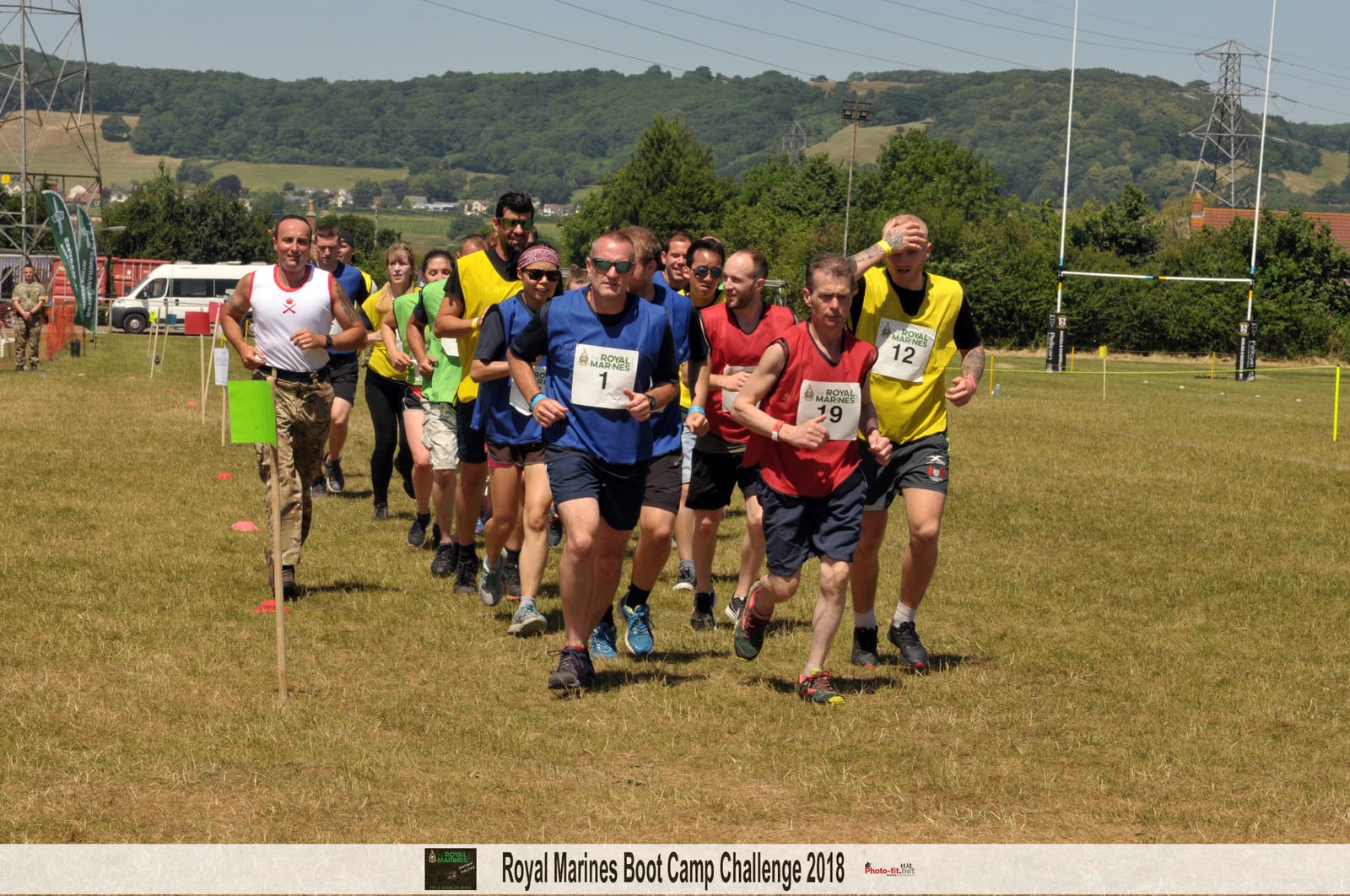 A group of people are running outdoors on a grassy field during the Royal Marines Boot Camp Challenge 2018. Each participant is wearing a numbered bib. In the background, there are hills, power lines, and trees under a clear sky.