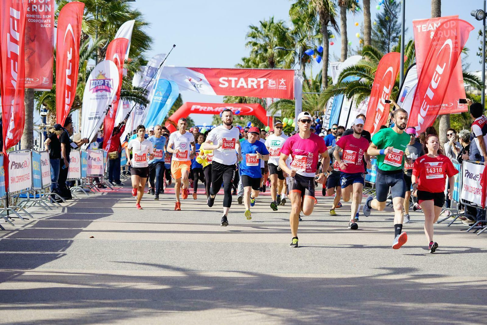 Runners participating in a marathon event, captured mid-race on a sunny day. They are surrounded by colorful banners and flags at the starting line, with palm trees and spectators lining the course. Many runners are wearing numbered bibs and athletic gear.