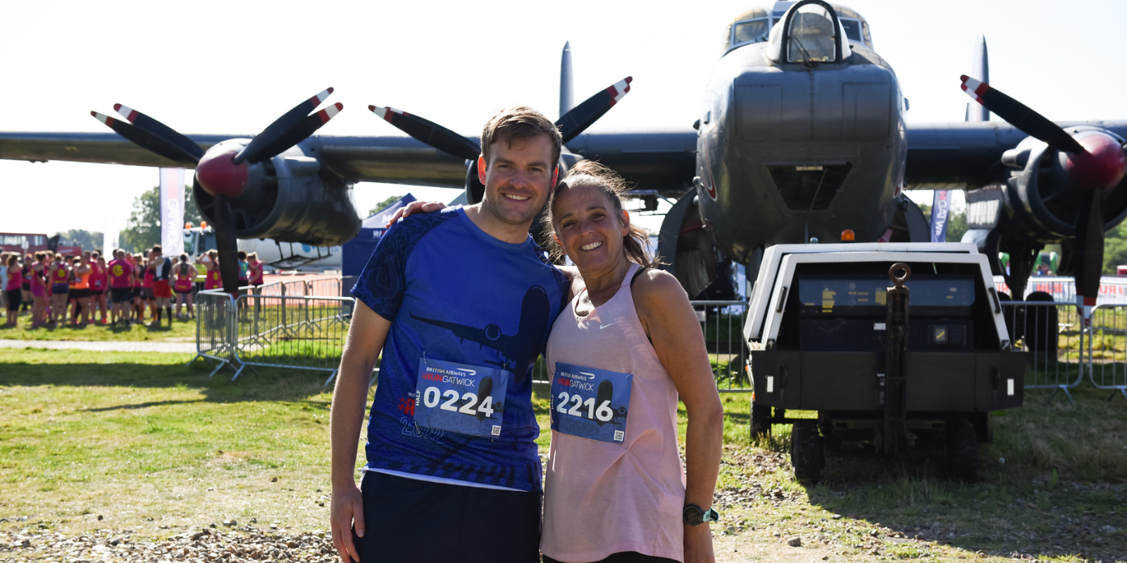 Two people stand smiling in front of a vintage aircraft with four propellers. Both wear race bibs, with numbers 0224 and 2216, suggesting participation in a running event. Other participants and spectators can be seen in the background.