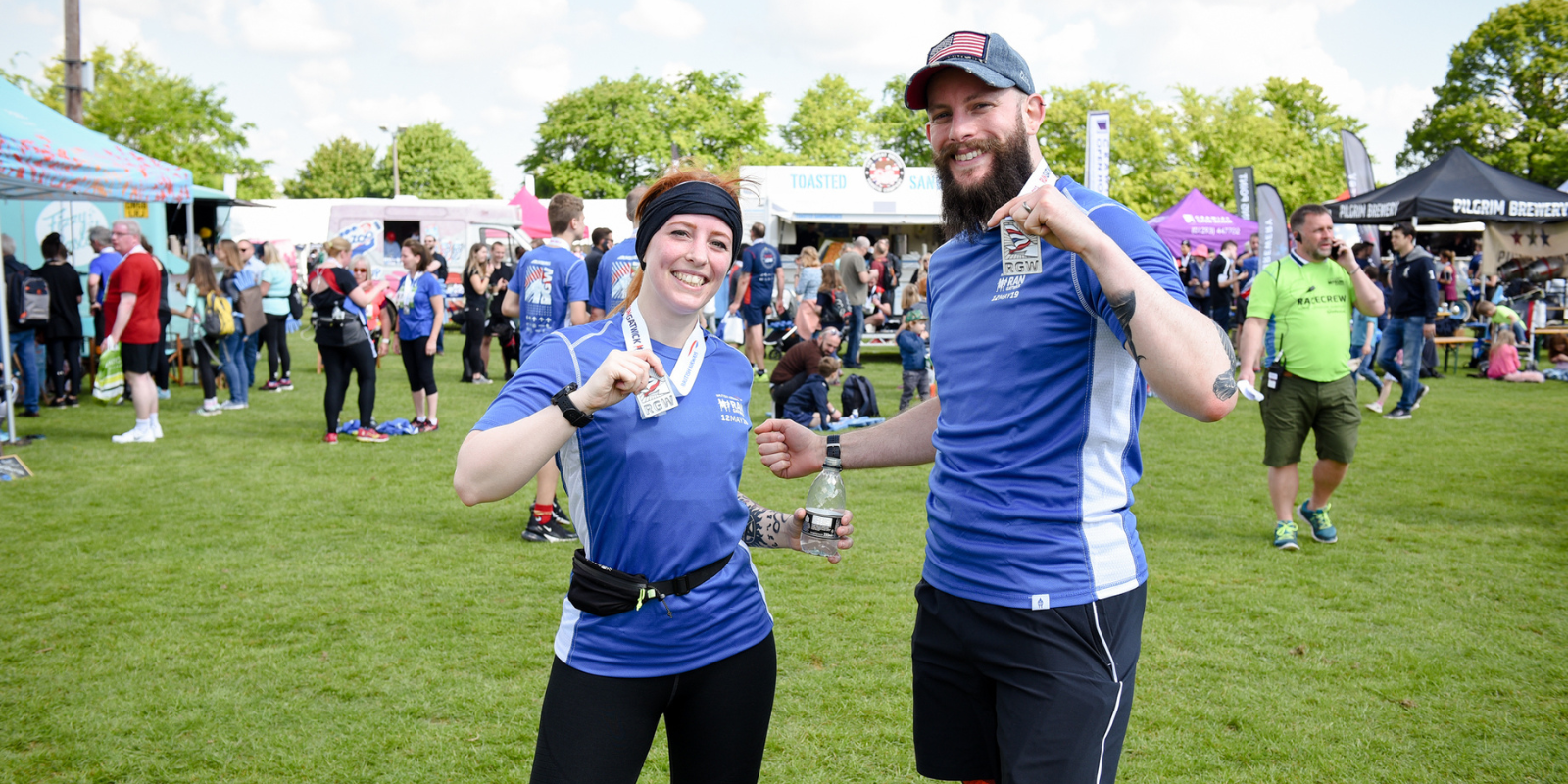 Two people in athletic gear celebrate with medals at a post-race event on a grassy field. They smile and pose with their medals, surrounded by other participants, tents, and a vibrant crowd on a sunny day.
