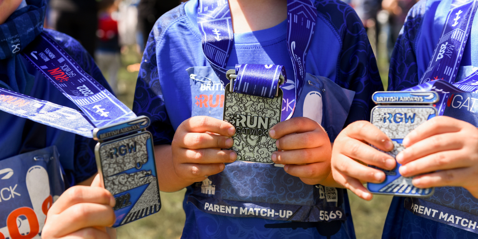 Three individuals wearing blue shirts and race bibs proudly display their medals after participating in British Airways' "Run Gatwick" event. Their shirts and medals bear the event's branding, and the medals are held up close to the camera for emphasis.