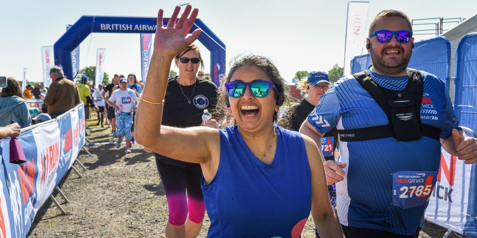 Runners smile and wave as they pass under a "British Airways" arch at an outdoor race event. A woman in a blue sleeveless top waves enthusiastically, and a man in a blue shirt with a race number jogs beside her. Other participants and spectators are visible in the background.