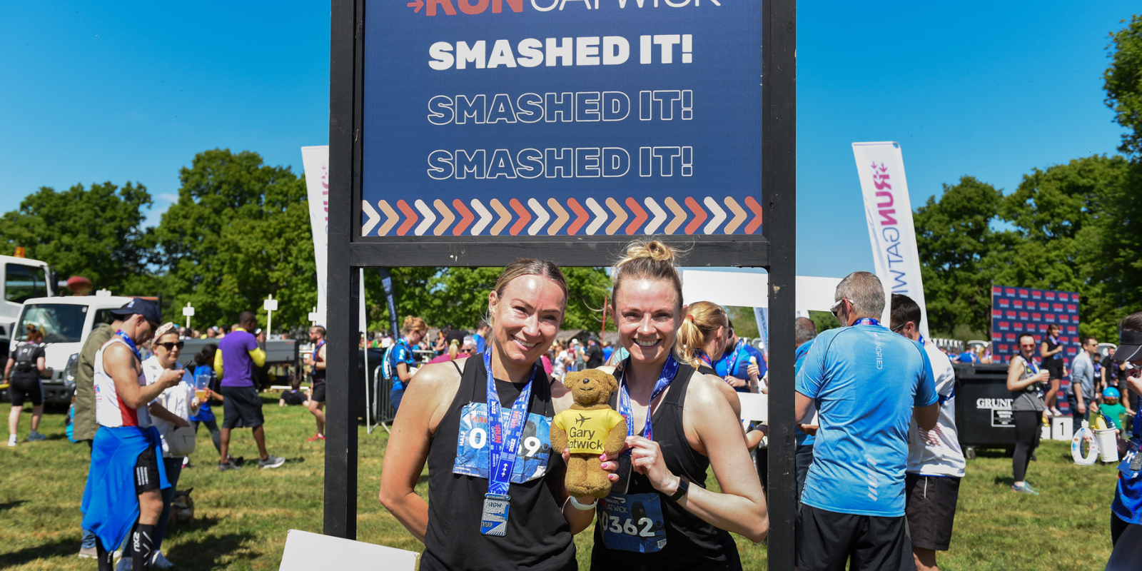 Two smiling women wearing race medals stand in front of a sign that reads "Smash It! Smashed It! Smashed It!" They are holding a brown teddy bear with a shirt that says, "Terry Tarry Canzian." Other runners are visible in the background at the outdoor event.