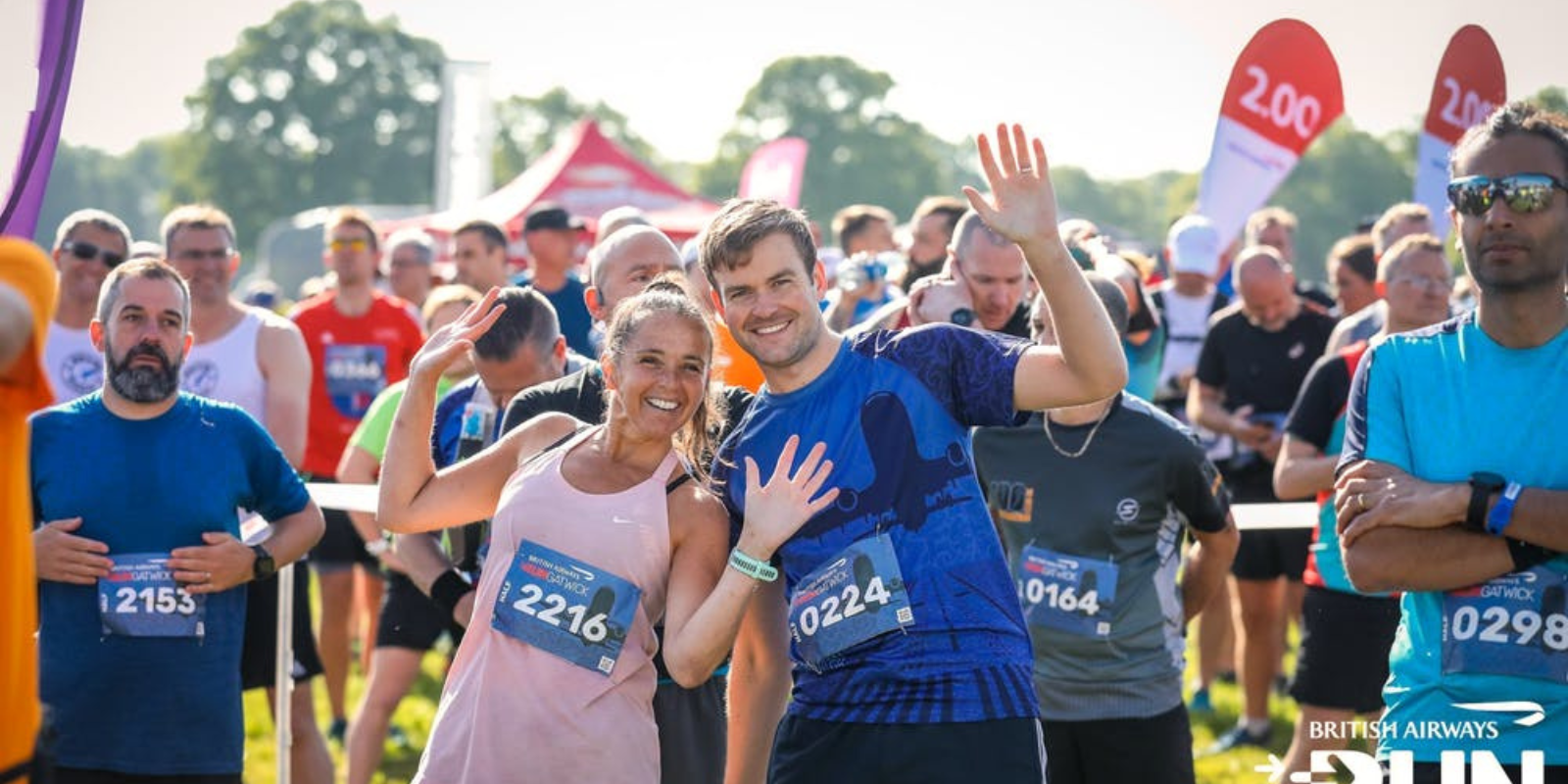A group of runners are gathered together outdoors at a starting line, smiling and waving at the camera. They are wearing race bibs. The background includes trees, event tents, and a banner that mentions "British Airways" and "Run.
