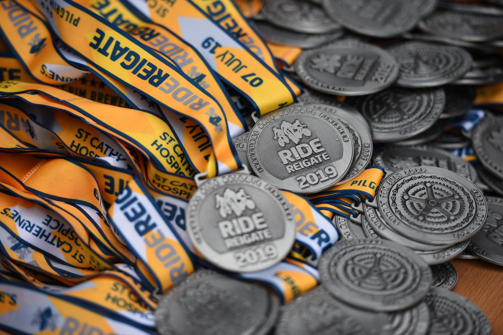 Close-up of a pile of medals from the Ride Reigate cycling event, dated 2019. The medals are silver with the event name and year etched on them, and are attached to yellow and blue ribbons that read "Ride Reigate" and "St. Catherine's Hospice.