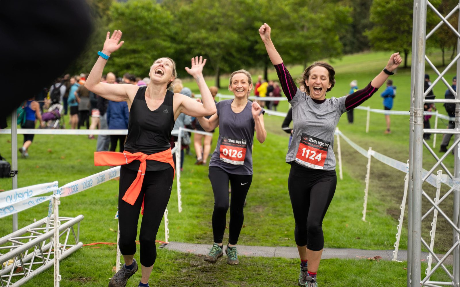 Three women runners, wearing race bibs numbered 0126 and 1124, cheer and raise their arms in celebration as they cross the finish line in an outdoor race. They are surrounded by greenery and other participants in the background.