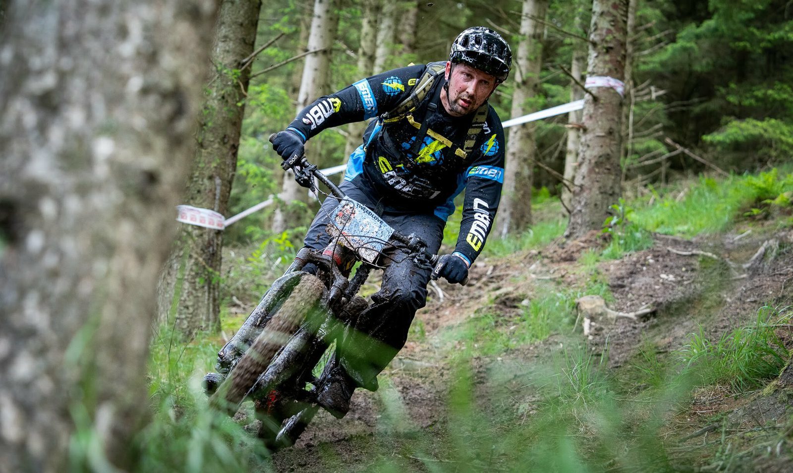 A cyclist wearing a black helmet and full-length cycling gear rides a mountain bike down a narrow, muddy forest trail. The cyclist is intensely focused, maneuvering through dense trees and greenery with caution tape visible in the background.