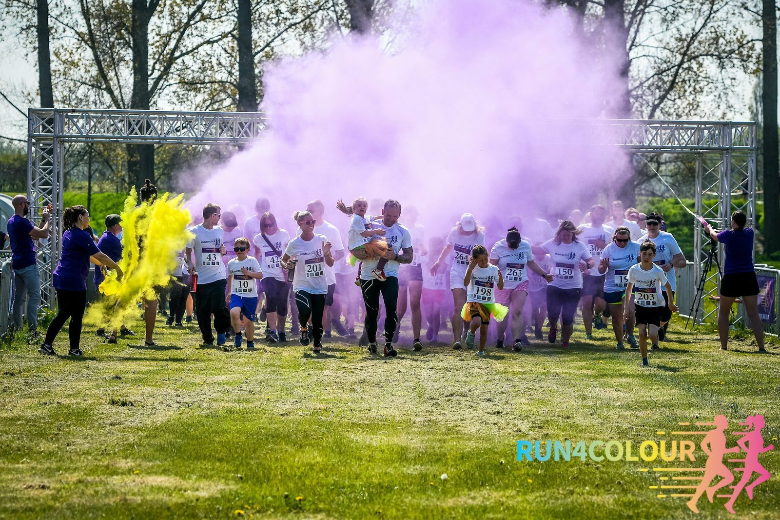Participants in a "Run 4 Colour" event are running through a purple and yellow cloud of color powder. They are wearing white shirts and numbered bibs. The event takes place outdoors with greenery and trees in the backdrop.
