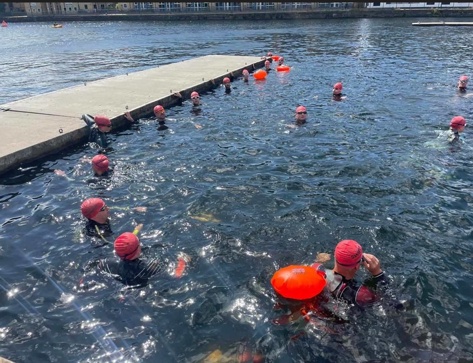 A group of people wearing red swim caps and wetsuits are swimming in a body of water next to a concrete dock. Some individuals are holding orange flotation devices while others are scattered around, treading water or preparing to swim.