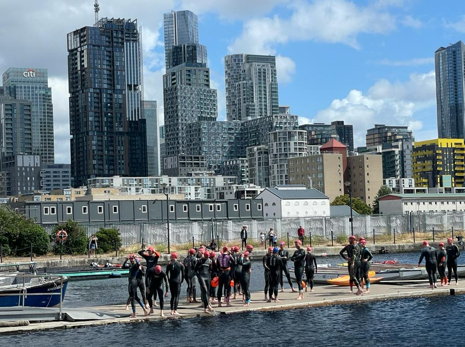 A group of athletes in wetsuits and red swim caps stand on a dock, preparing for a swim in a body of water. Skyscrapers and modern high-rise buildings form the urban background under a partly cloudy sky. Several kayaks and boats are visible nearby.