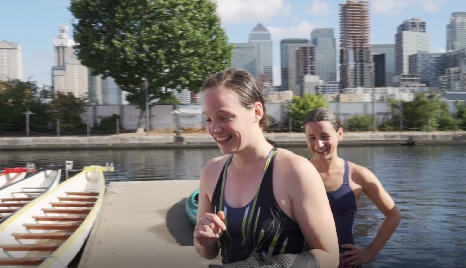 Two women are standing by the waterfront, smiling, and appearing to engage in post-rowing activities. One wears a black and green striped athletic top, while the other wears a black outfit. A docked rowing boat and a cityscape with tall buildings are in the background.