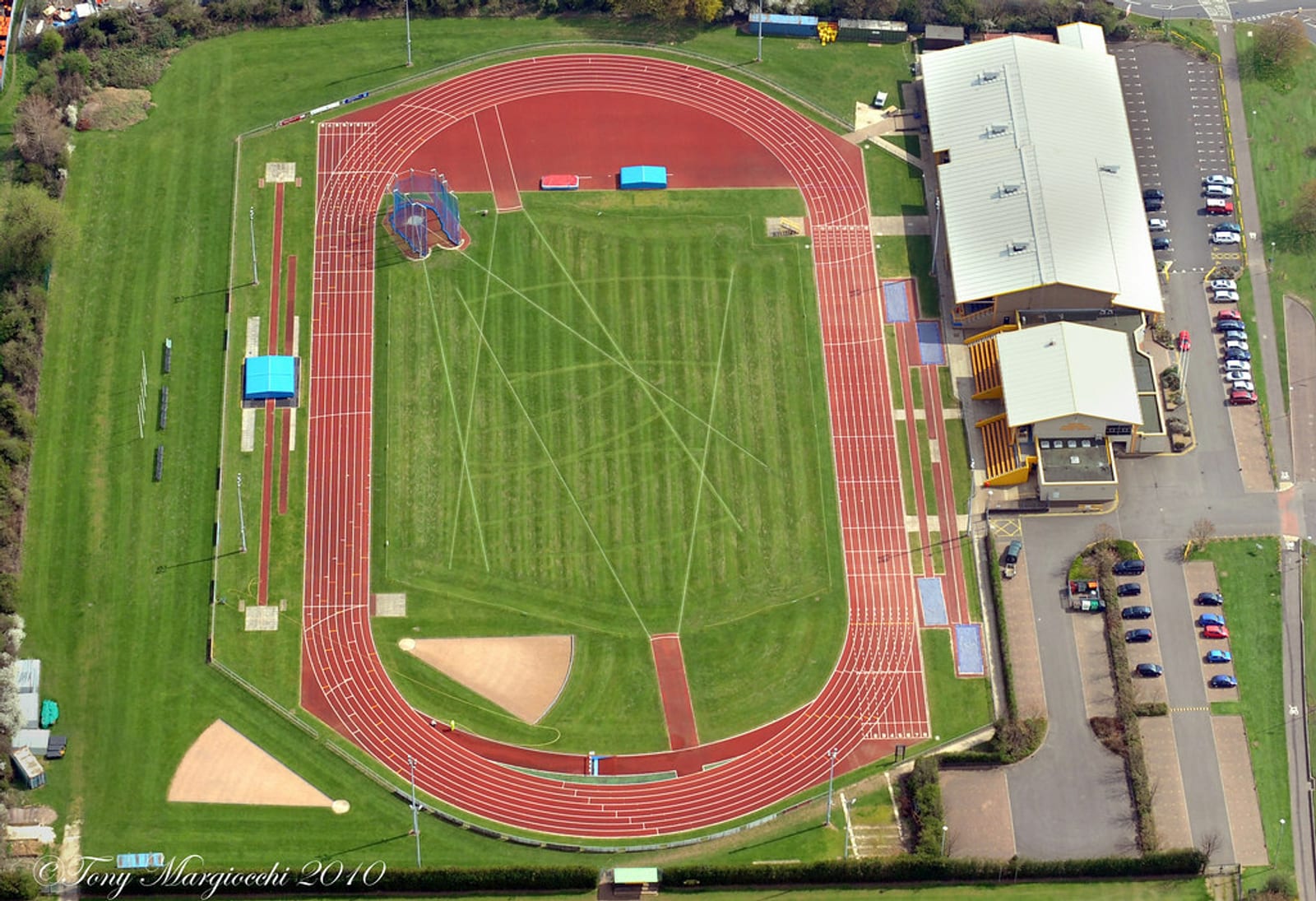 Aerial view of an athletics stadium featuring a red running track encircling a green infield with various field event areas. Adjacent to the track is a large building with a parking lot. The stadium is surrounded by trees and additional walkways.