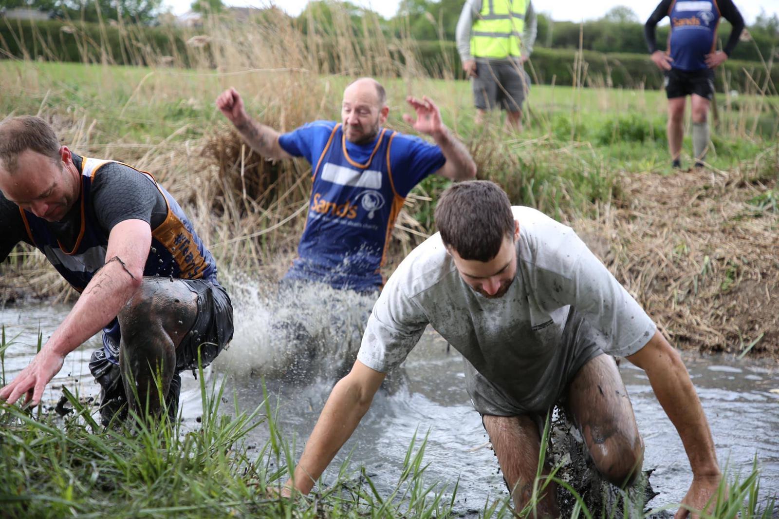 Three men are navigating through a muddy water obstacle during an outdoor event. One man in a blue shirt is splashing into the water, while the others, in gray shirts, are crawling out of the mud. A person in a reflective vest stands nearby.