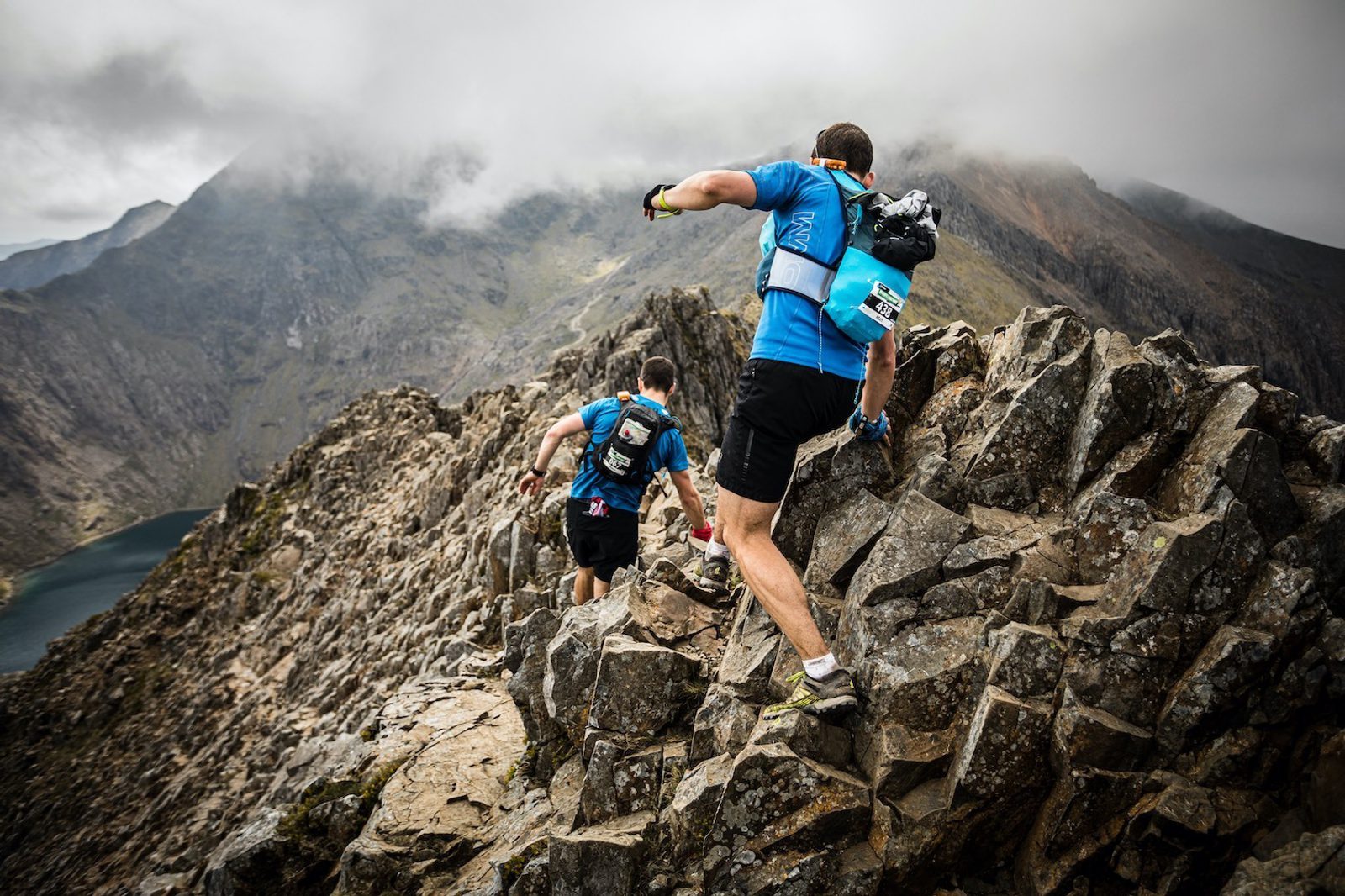 Two runners in blue shirts and black shorts navigate a rugged, rocky mountain ridge under cloudy skies. They are equipped with hydration packs and race bibs. A scenic view of mountains and a lake is visible in the background.