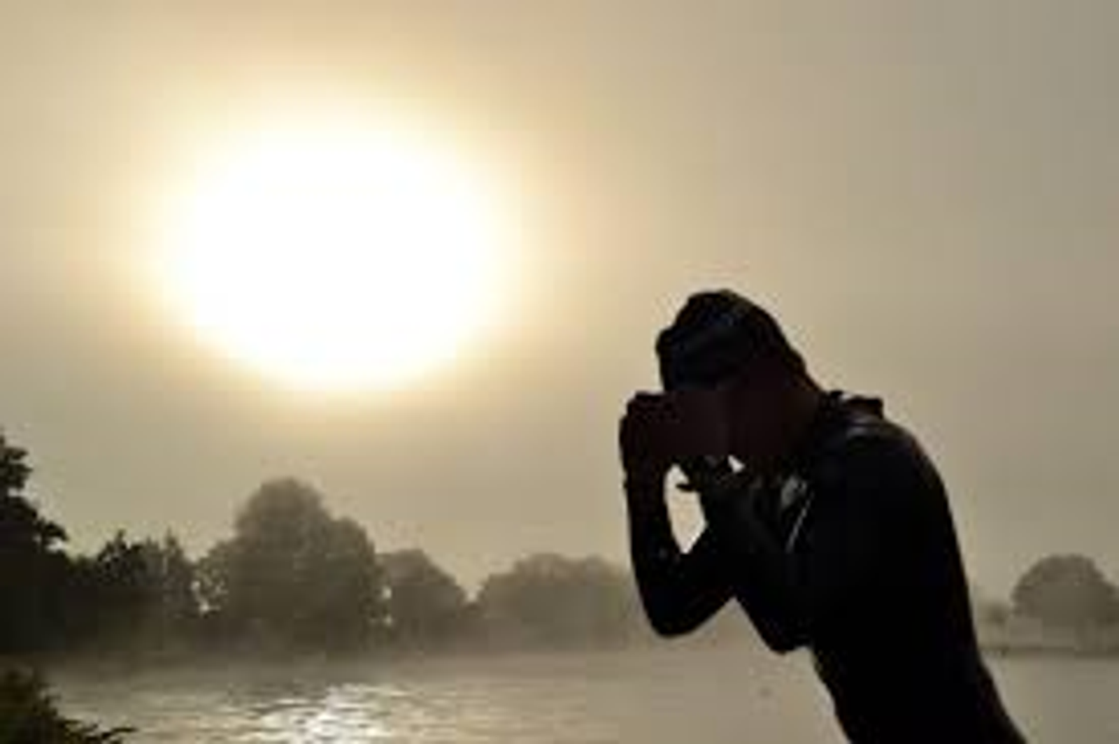 A person in a wetsuit and swim cap stands by a body of water with their hands held together near their face, appearing deep in thought or prayer. The sun is low in the sky, creating a bright glow and casting long shadows on the calm water surface.