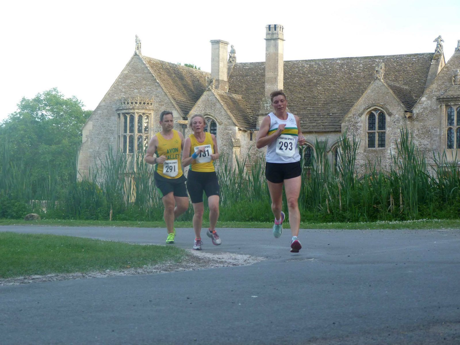 Three runners in athletic gear, wearing bibs 291, 175, and 293, running on a paved path. A historic stone building with tall, narrow windows and chimneys is in the background, surrounded by lush greenery. The sky is clear.