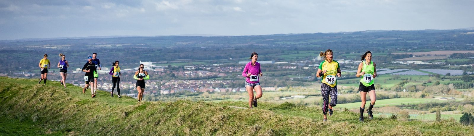People running a race on a grassy hillside with a scenic countryside view in the background. The runners are wearing numbered bibs and colorful athletic gear under a partly cloudy sky.